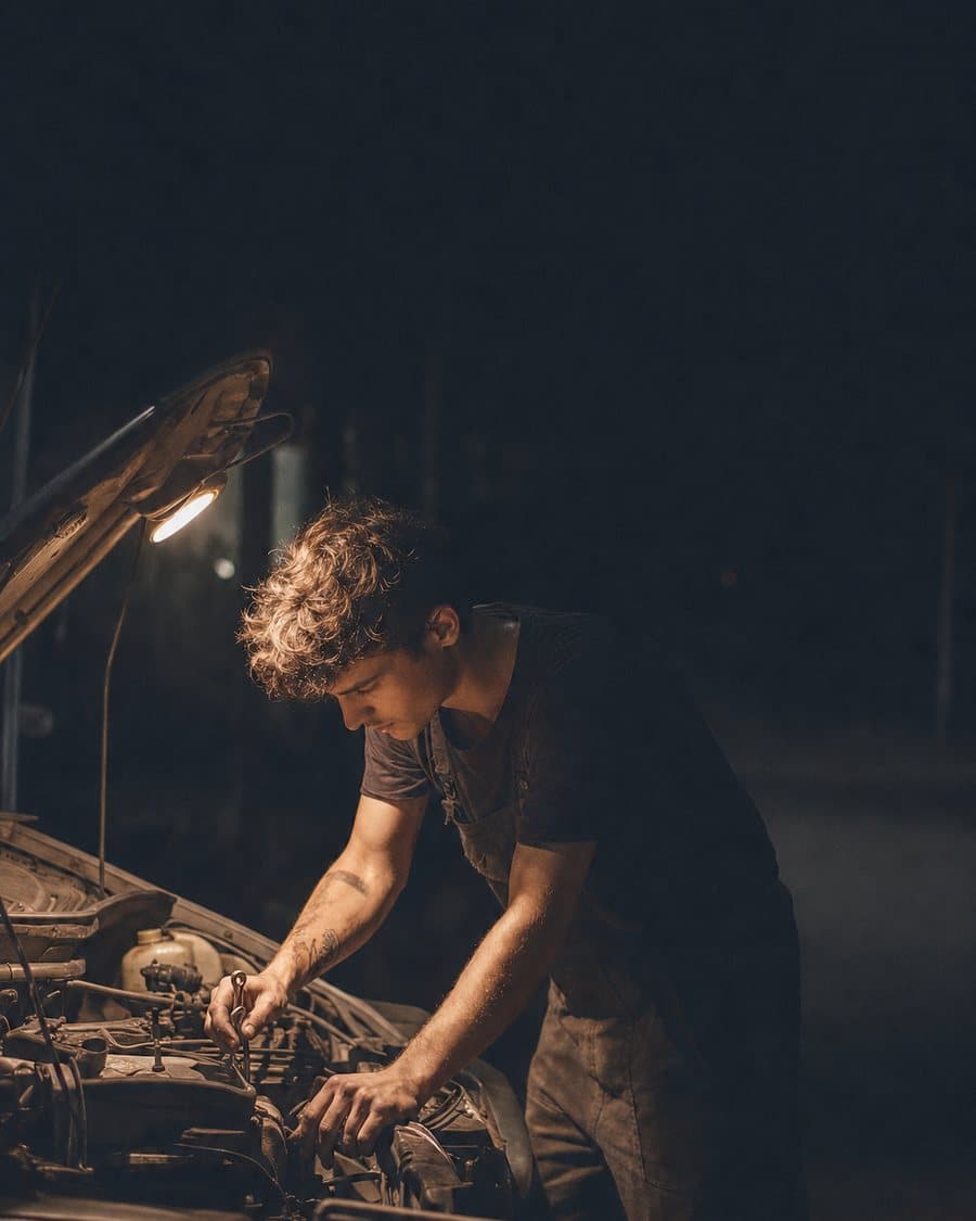 A mechanic leaning into an engine bay under a work light at dusk